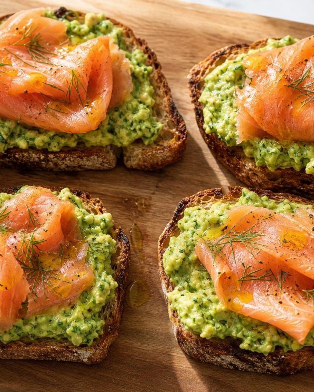 Four pieces of avocado toast topped with smoked salmon and fresh dill on a wooden board