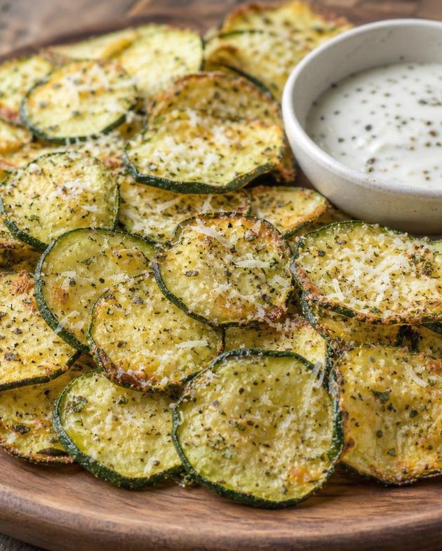 Golden crispy baked zucchini chips sprinkled with parmesan and Italian herbs on a wooden platter