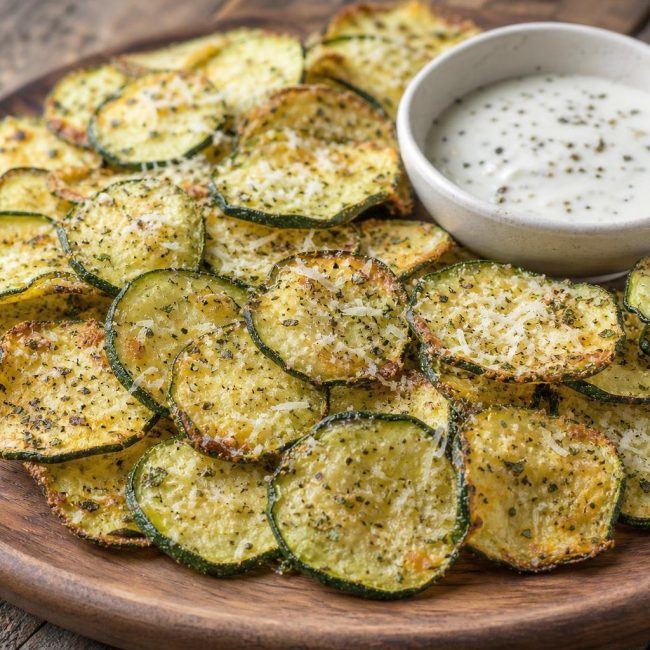 Golden crispy baked zucchini chips sprinkled with parmesan and Italian herbs on a wooden platter