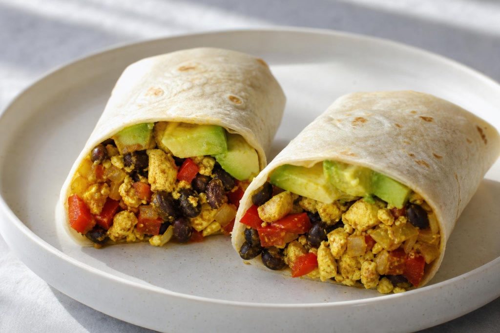 Two breakfast burrito halves filled with tofu scramble, black beans, red bell pepper, and avocado on a white plate.