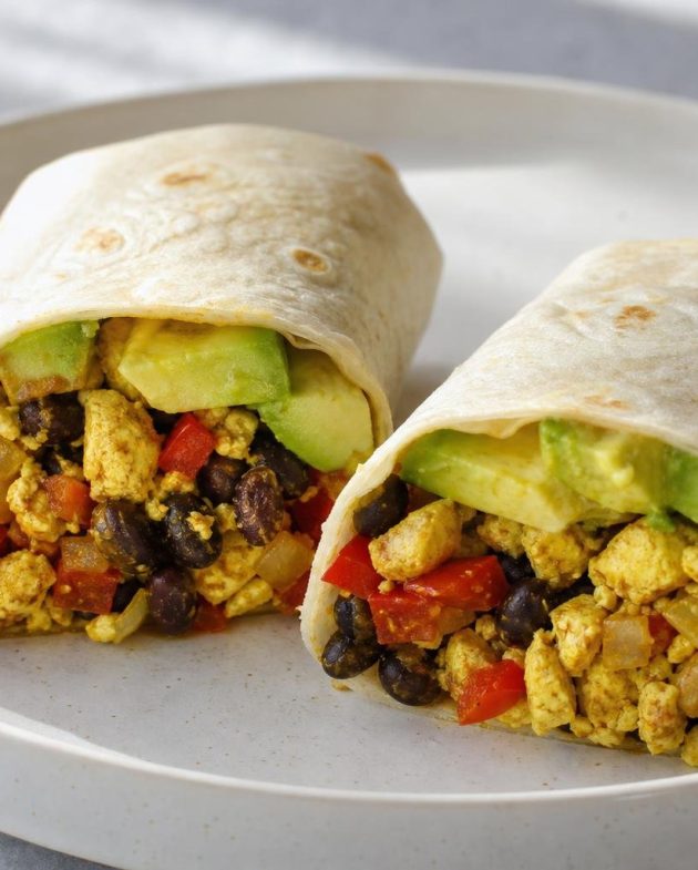 Two breakfast burrito halves filled with tofu scramble, black beans, red bell pepper, and avocado on a white plate.