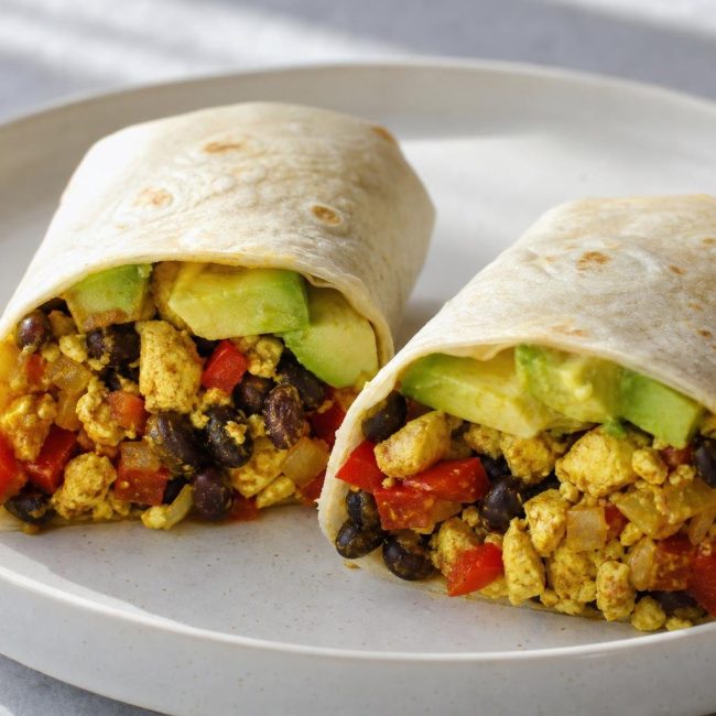 Two breakfast burrito halves filled with tofu scramble, black beans, red bell pepper, and avocado on a white plate.