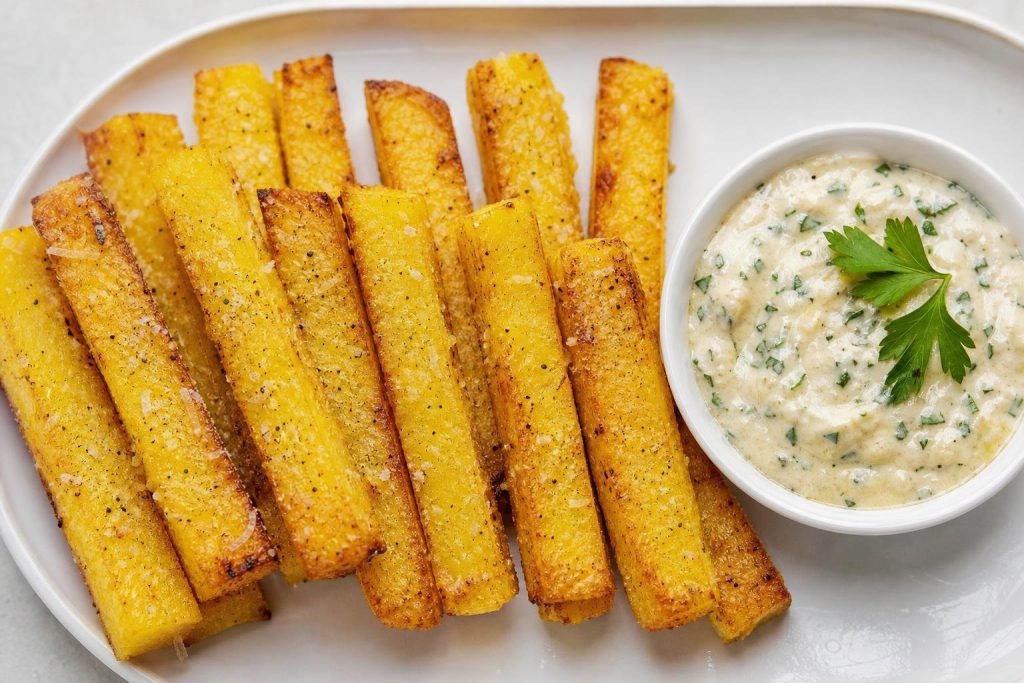 Crispy baked polenta fries served with a creamy garlic herb aioli in a bowl on the side.