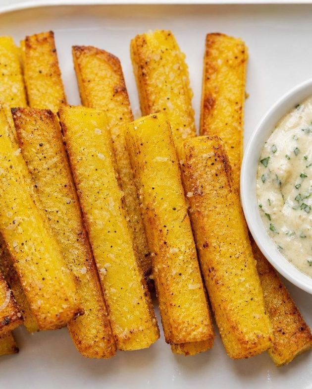 Crispy baked polenta fries served with a creamy garlic herb aioli in a bowl on the side.