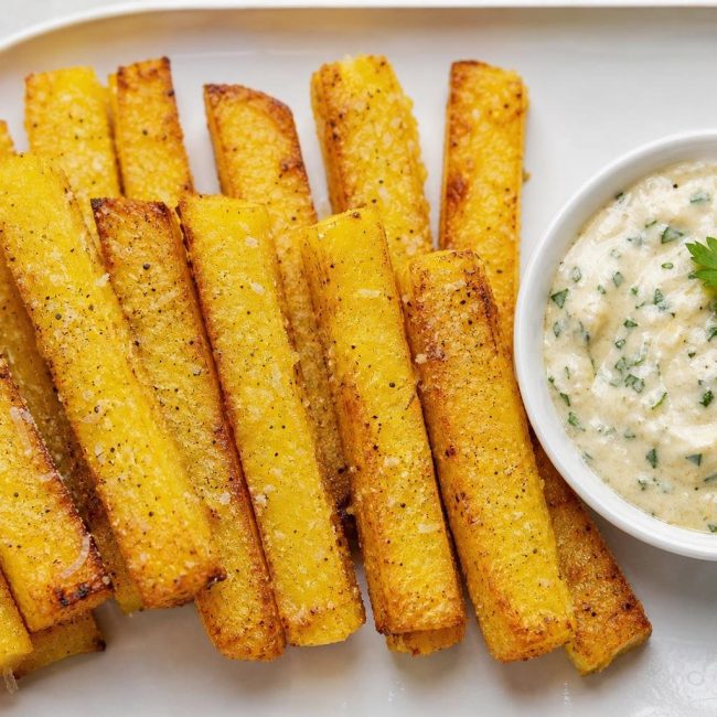 Crispy baked polenta fries served with a creamy garlic herb aioli in a bowl on the side.