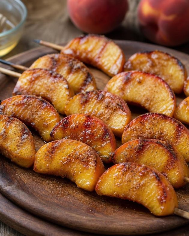 Grilled peach skewers brushed with honey and sprinkled with cinnamon sugar on a wooden platter with visible grill marks.