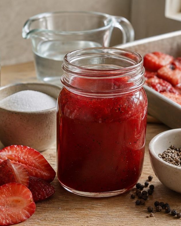 Jar of roasted strawberry and black pepper shrub with fresh strawberries and cracked black pepper on wooden surface.
