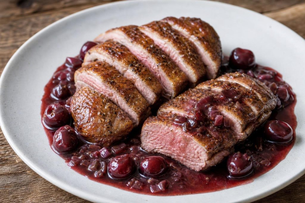Sliced seared duck breast with cherry-port sauce and thyme garnish on white plate.
