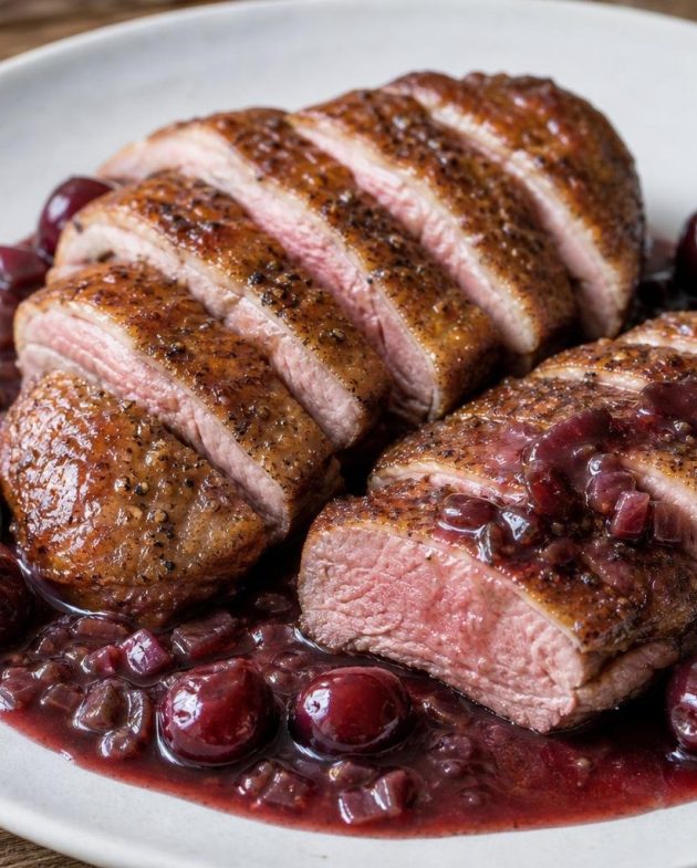 Sliced seared duck breast with cherry-port sauce and thyme garnish on white plate.