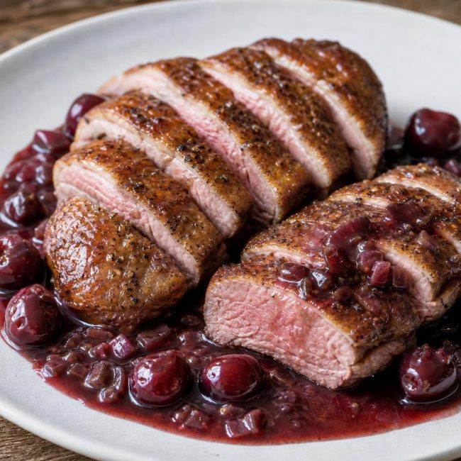 Sliced seared duck breast with cherry-port sauce and thyme garnish on white plate.